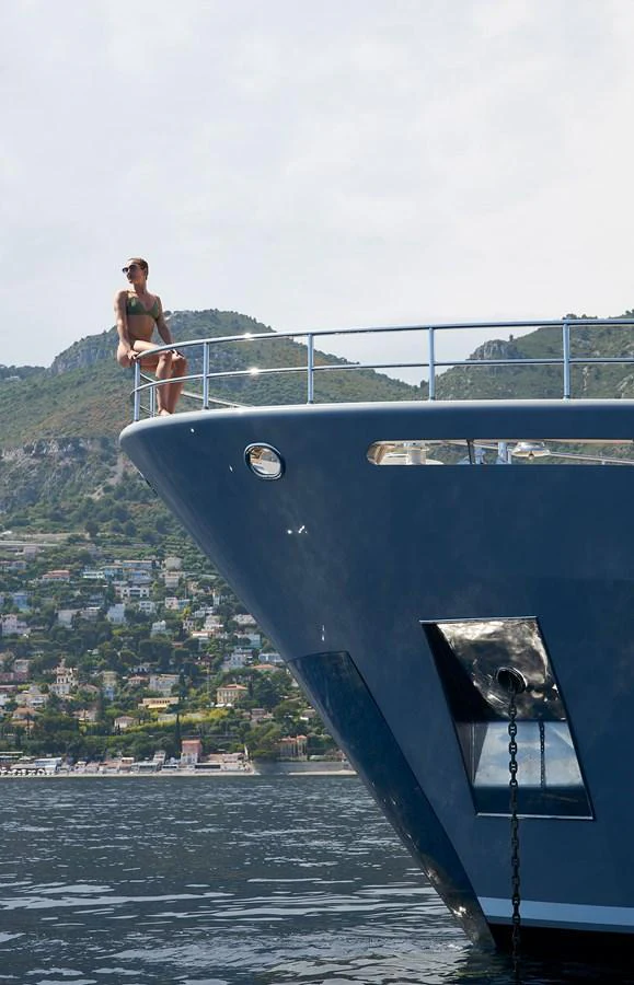A person enjoys the view from the bow of a sleek yacht against a scenic coastal backdrop. aboard GALENA Yacht for Sale