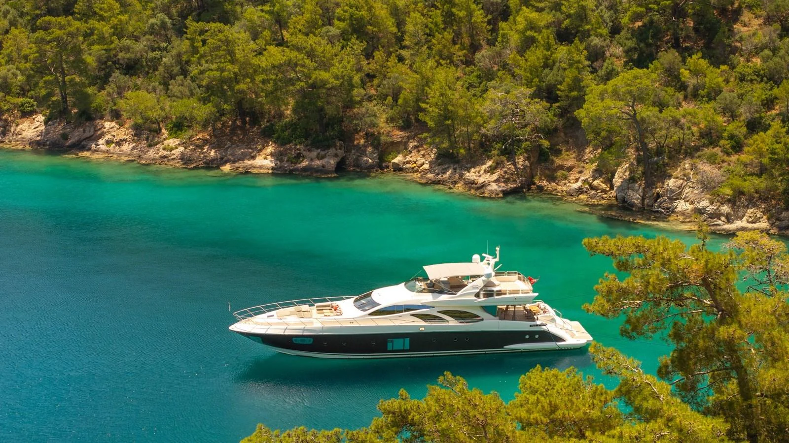 A sleek yacht is anchored in a serene, turquoise bay surrounded by lush greenery. aboard EROSSEA Yacht for Sale