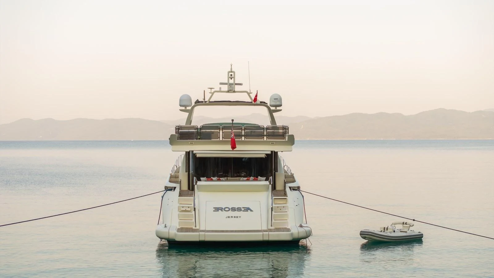 The yacht is anchored in calm waters, showcasing its elegant rear profile with a tender nearby. aboard EROSSEA Yacht for Sale