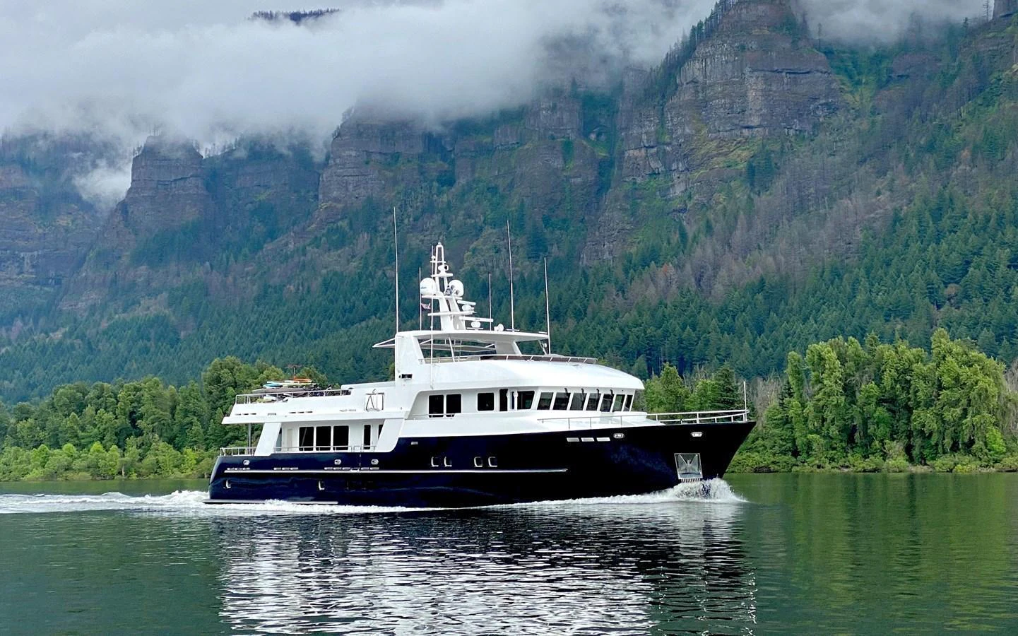 A sleek yacht cruises through calm waters with a lush, mountainous backdrop. aboard SAMSARA Yacht for Sale