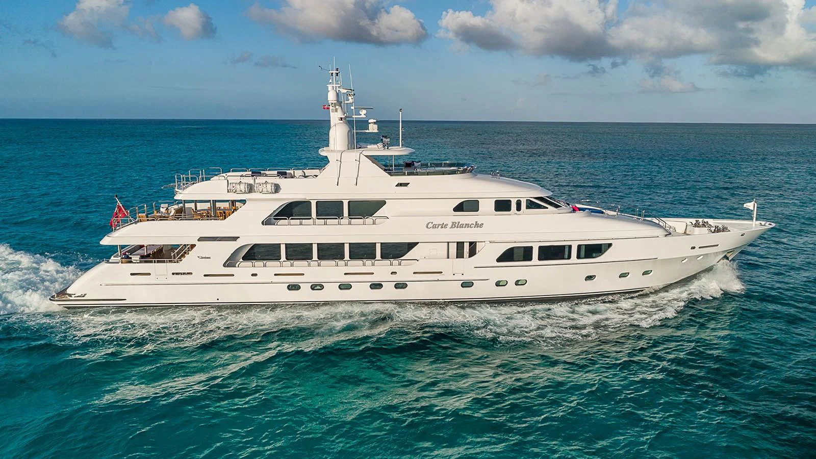 A luxurious yacht cruises through clear blue waters under a partly cloudy sky. aboard CARTE BLANCHE Yacht for Sale