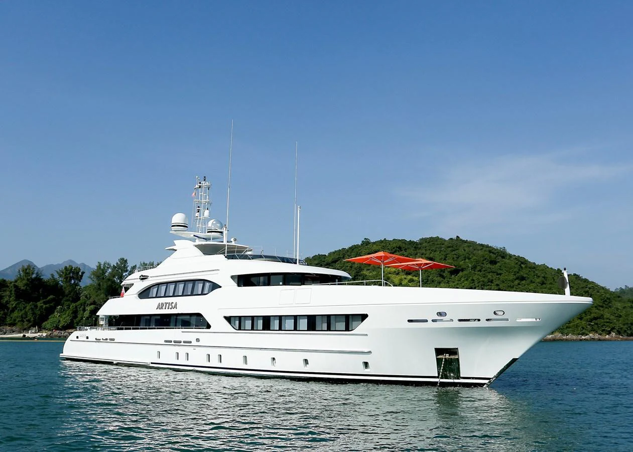 A sleek white yacht glides gracefully on calm waters with a scenic backdrop of lush green hills. aboard HEESEN 47M Yacht for Sale