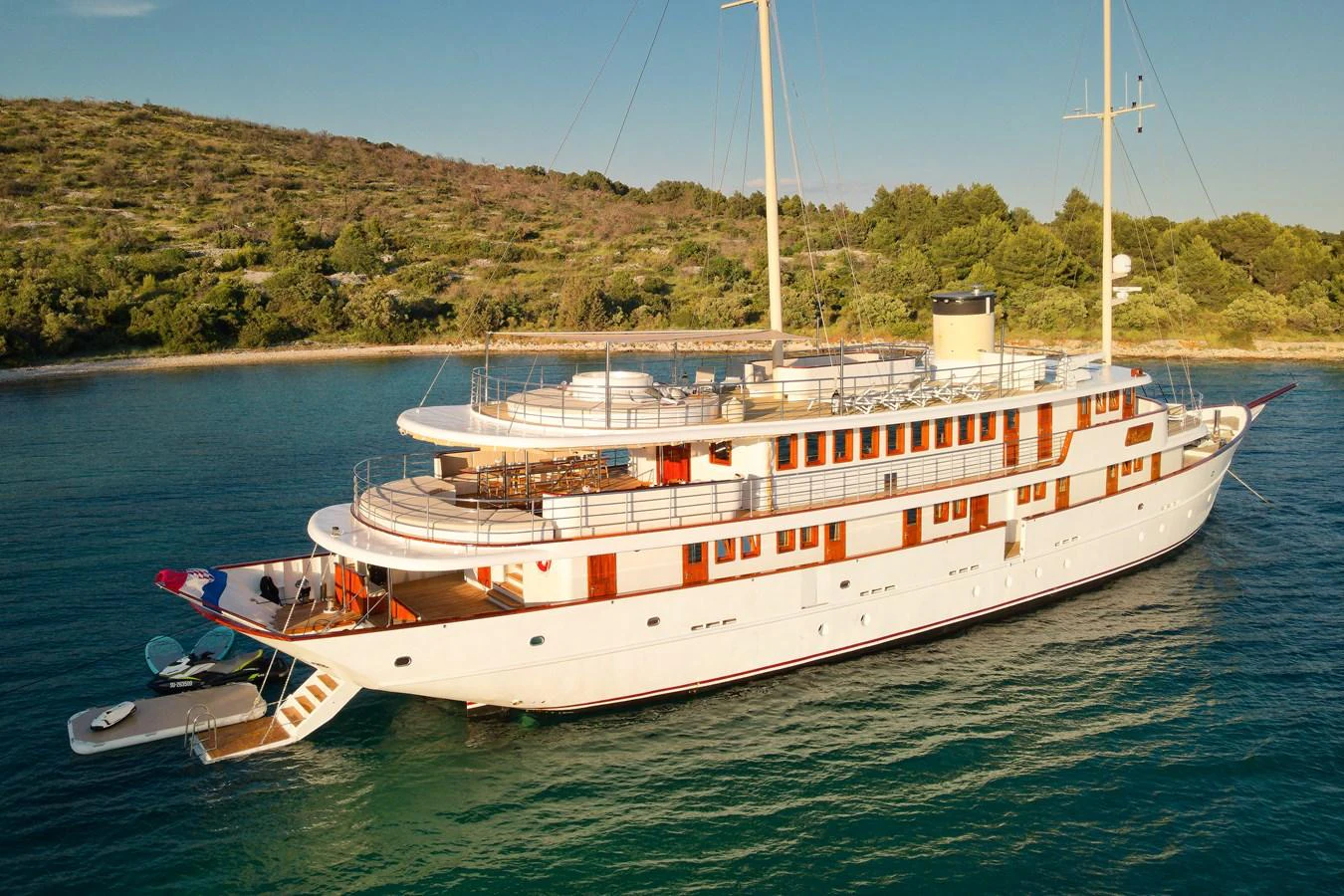 A classic yacht with elegant lines is anchored near a lush, green coastline. aboard BELLEZZA Yacht for Sale