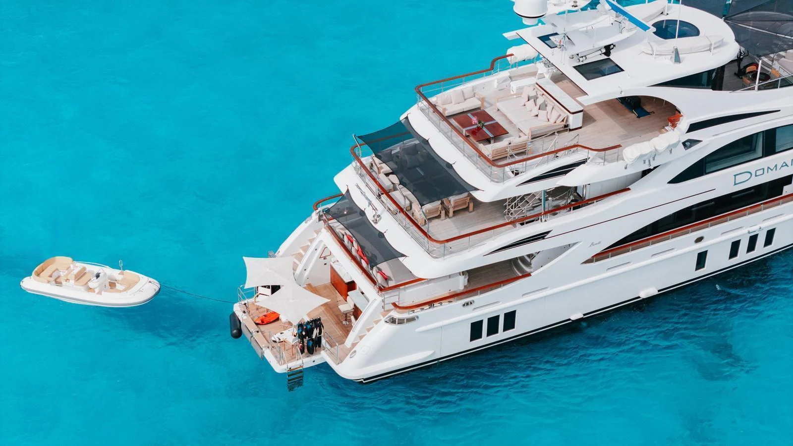 A luxurious yacht is anchored in crystal-clear blue waters, showcasing its expansive decks and a tender boat nearby. aboard DOMANI Yacht for Sale