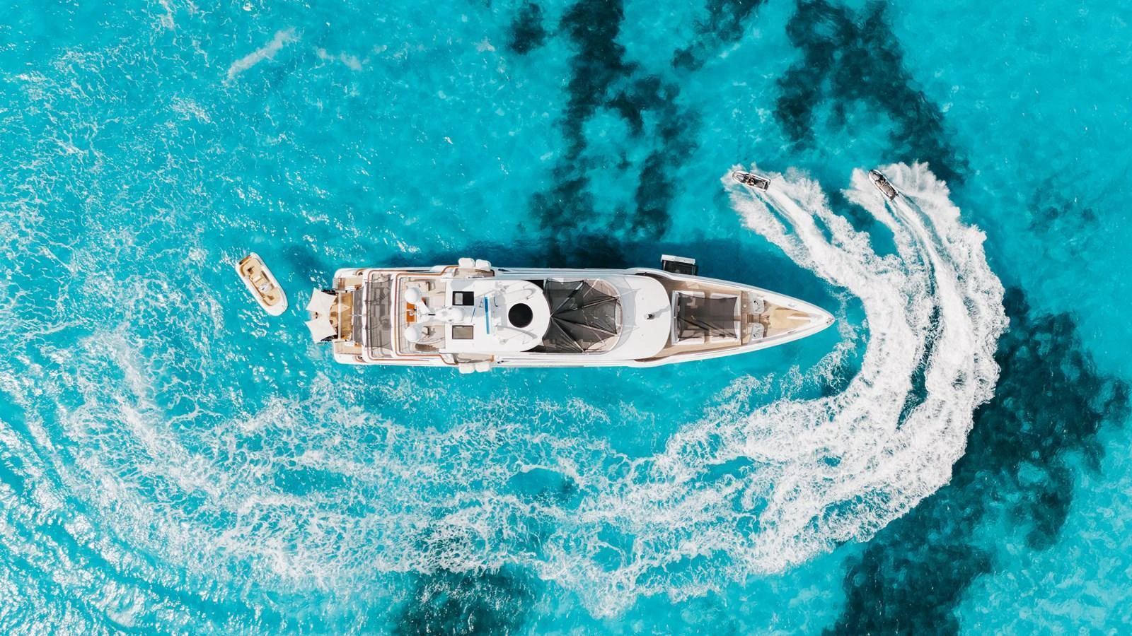 A stunning aerial view of a yacht cruising through crystal-clear turquoise waters, accompanied by a tender and a jet ski. aboard DOMANI Yacht for Sale