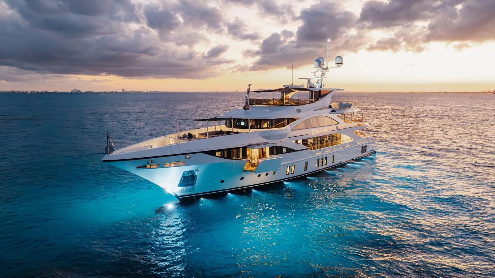 A stunning yacht is illuminated against a serene ocean backdrop at sunset. aboard DOMANI Yacht for Sale
