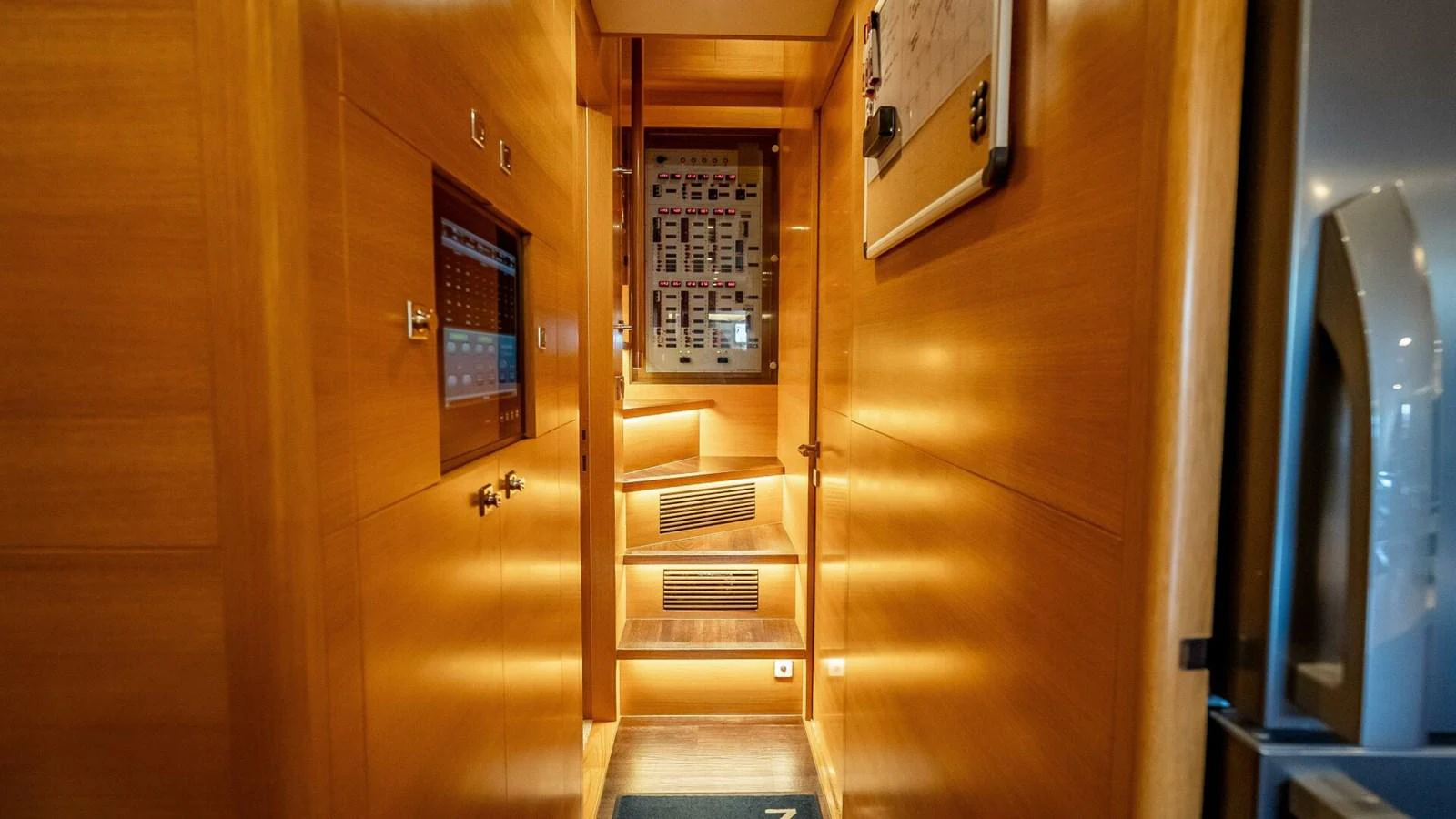 A warmly lit hallway with wooden paneling and a control panel, leading to a staircase. aboard SHORT STORY Yacht for Sale