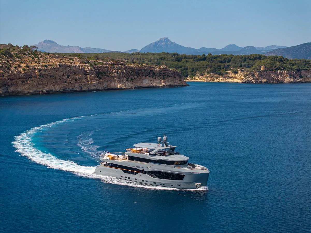 A luxurious yacht cruises through pristine blue waters with a scenic coastline in the background. aboard OSCAR Yacht for Sale