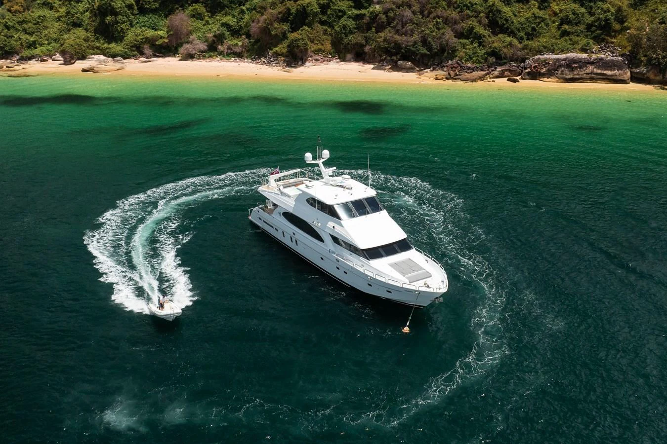 A luxurious yacht is anchored in a serene, emerald-green bay with a tender creating a circular wake around it. aboard SASSY Yacht for Sale