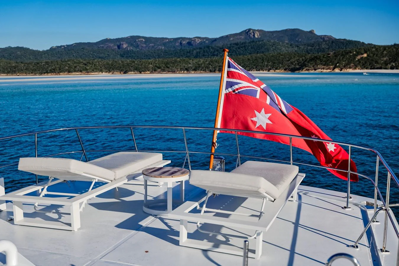 The sundeck offers a serene view of the coastline, featuring comfortable lounge chairs and a vibrant flag waving in the breeze. aboard VICTORIA Yacht for Sale