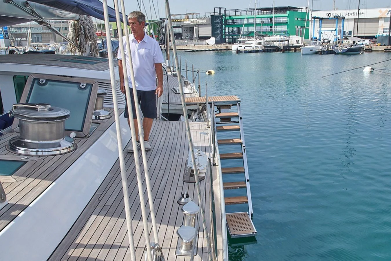 A man stands on the deck of a yacht docked at a marina, showcasing the vessel's sleek design and polished woodwork. aboard CYCLOS III Yacht for Sale