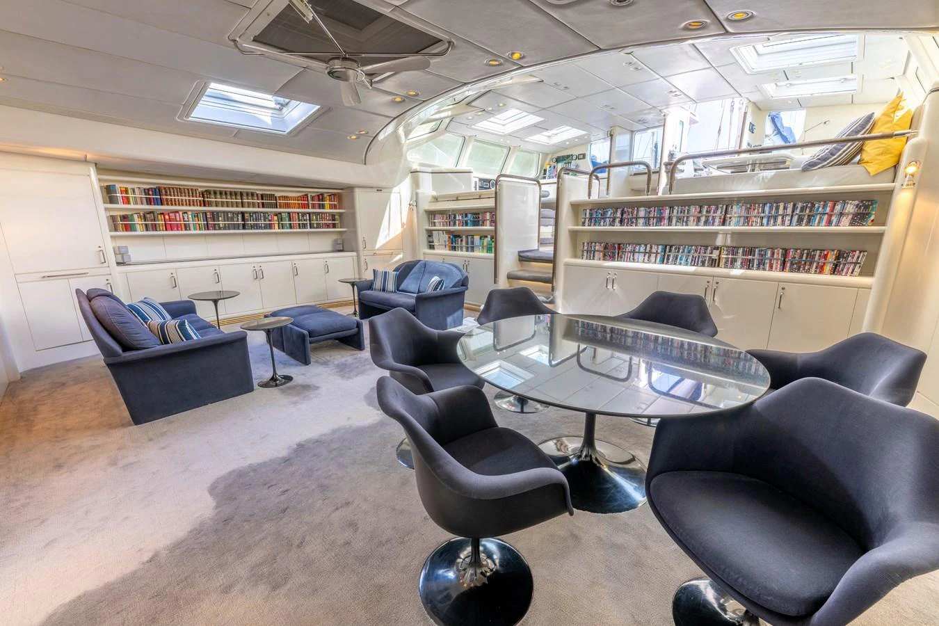The yacht's salon features a cozy seating area with a library and a dining table, bathed in natural light from overhead skylights. aboard CYCLOS III Yacht for Sale