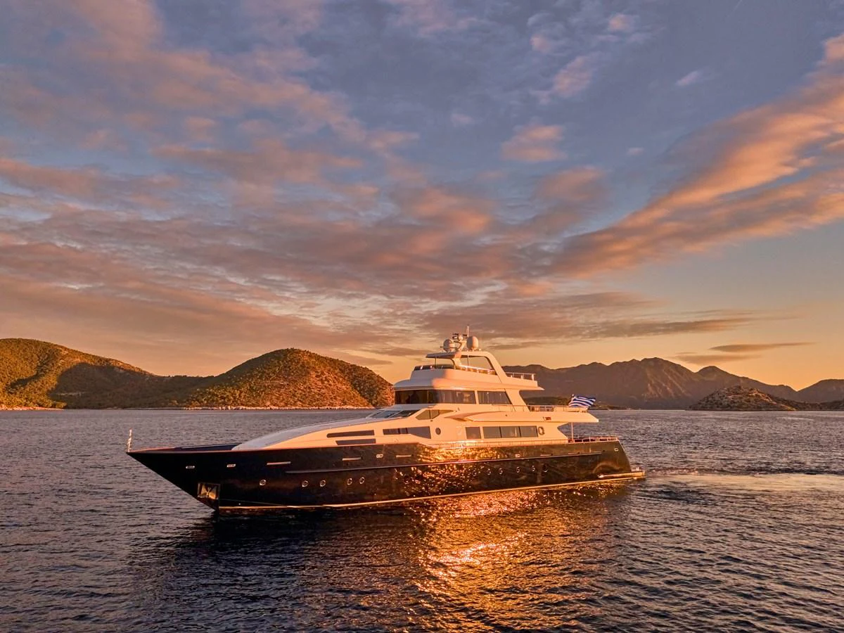 A stunning yacht glides through calm waters against a backdrop of picturesque hills and a vibrant sunset sky. aboard CONDOR A Yacht for Sale