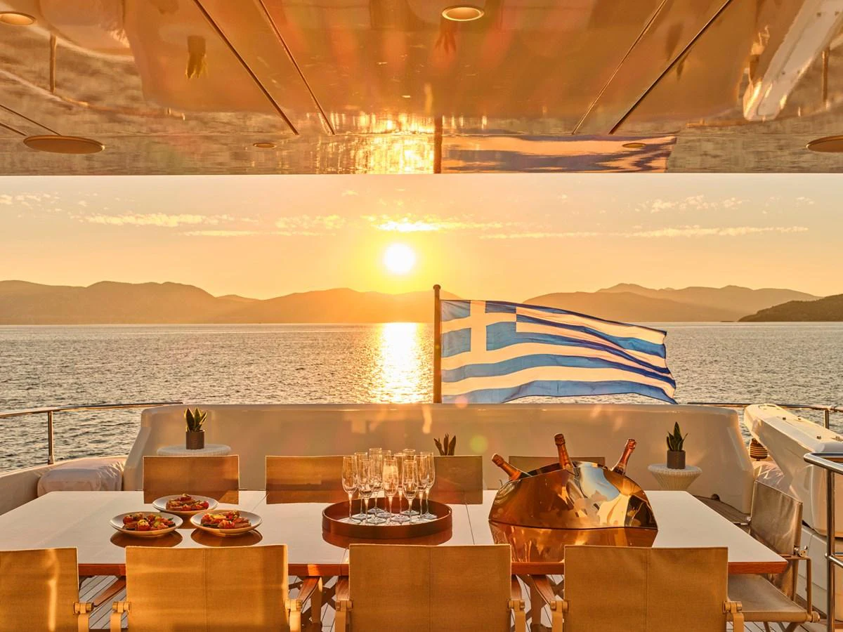 The aft deck offers a stunning sunset view with a dining setup and a Greek flag waving in the breeze. aboard CONDOR A Yacht for Sale