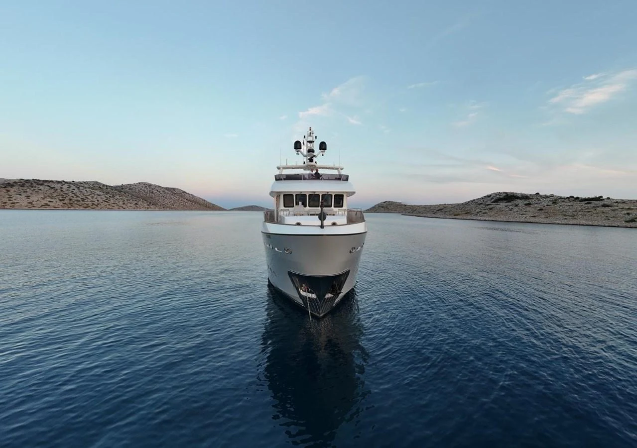 The yacht is anchored in a serene bay, showcasing its elegant bow against a backdrop of calm waters and distant hills. aboard GRANIL Yacht for Sale