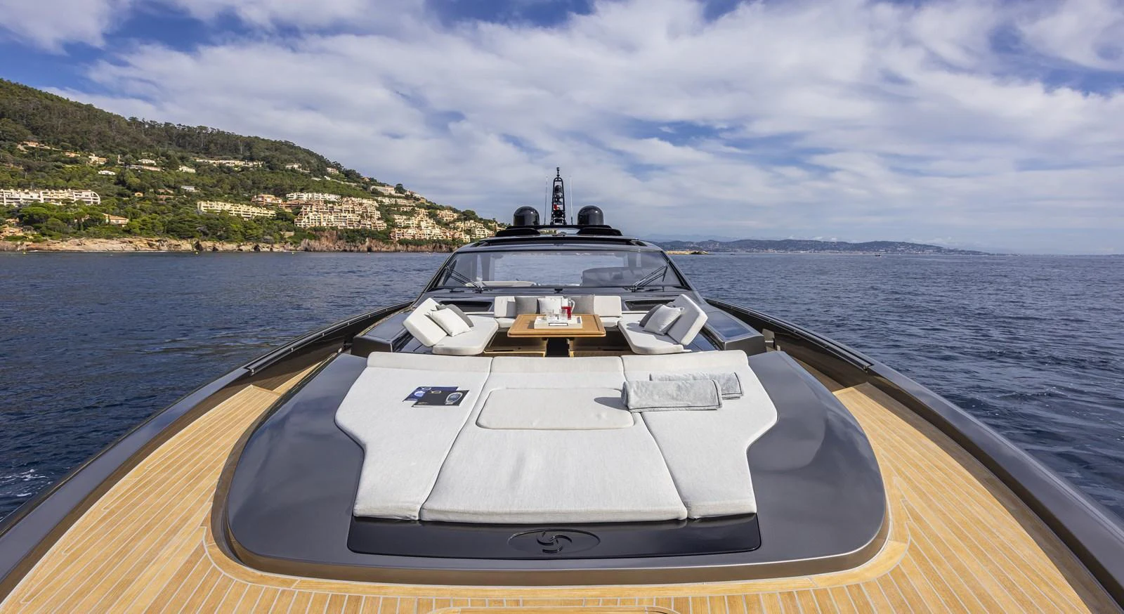 The yacht's foredeck features a luxurious sunbathing area with plush cushions and a central table, set against a scenic coastal backdrop. aboard SEXY ME Yacht for Sale