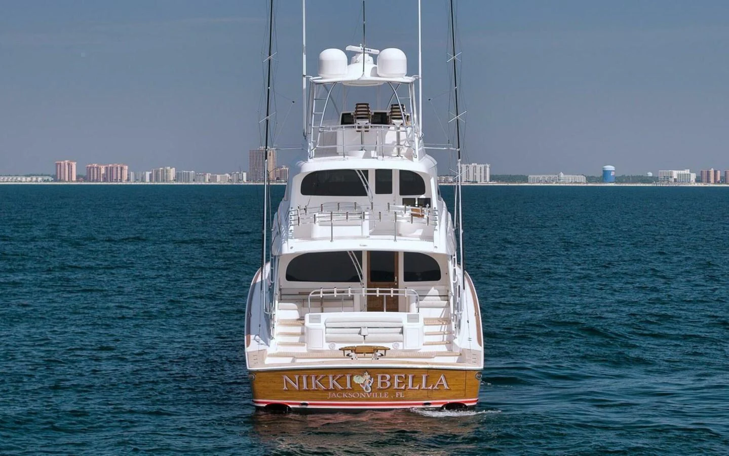The yacht is seen from the rear, showcasing its elegant design and spacious deck against a coastal backdrop. aboard NIKKI BELLA Yacht for Sale