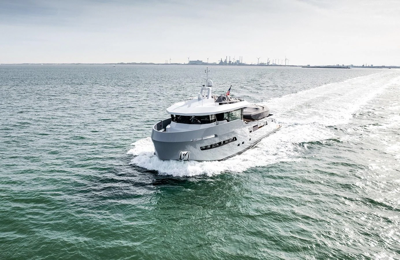 A sleek yacht cruises through calm waters with an industrial skyline in the background. aboard 27 CROSSOVER Yacht for Sale