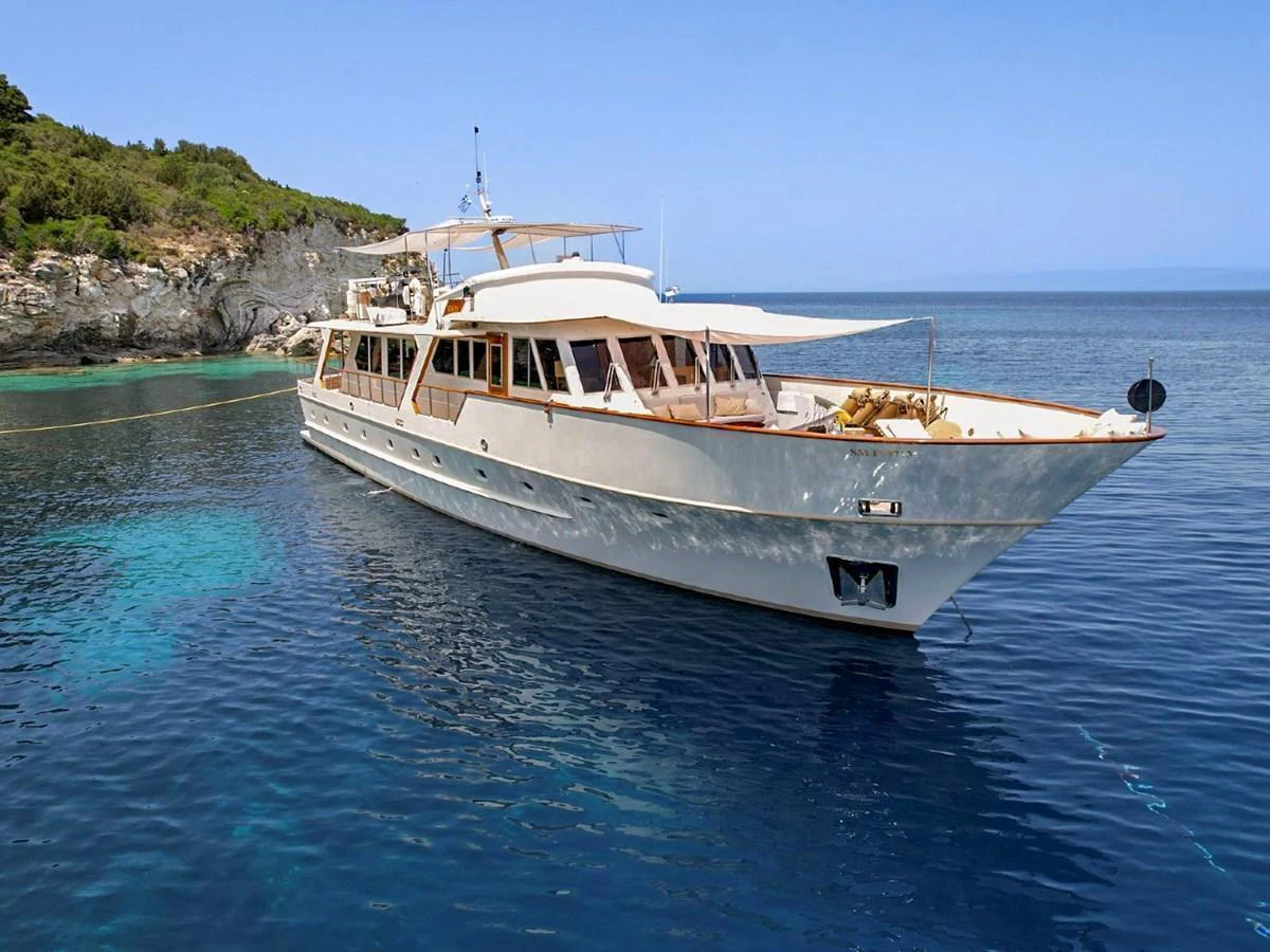 A classic yacht is anchored in a serene coastal area with clear blue waters and lush greenery in the background. aboard STALCA Yacht for Sale