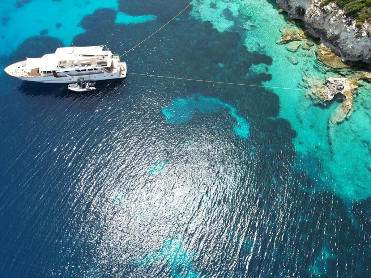 A luxurious yacht is anchored in crystal-clear turquoise waters near a rocky coastline. aboard STALCA Yacht for Sale