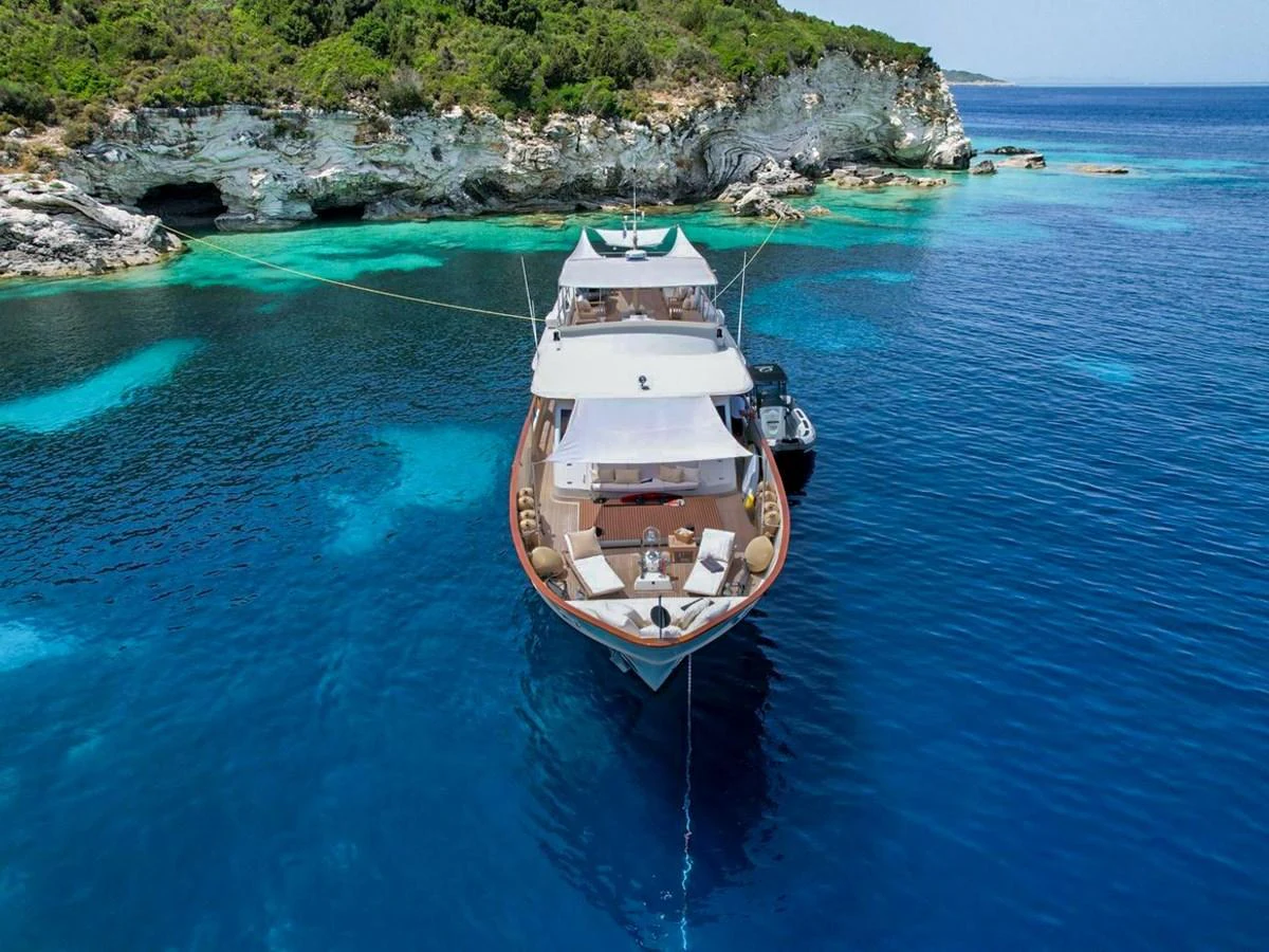 A luxurious yacht is anchored in a serene, turquoise bay with lush greenery and rocky cliffs in the background. aboard STALCA Yacht for Sale