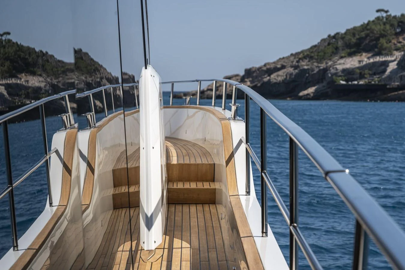 The yacht's bow features sleek wooden decking and polished railings, set against a serene coastal backdrop. aboard ORCA I Yacht for Sale