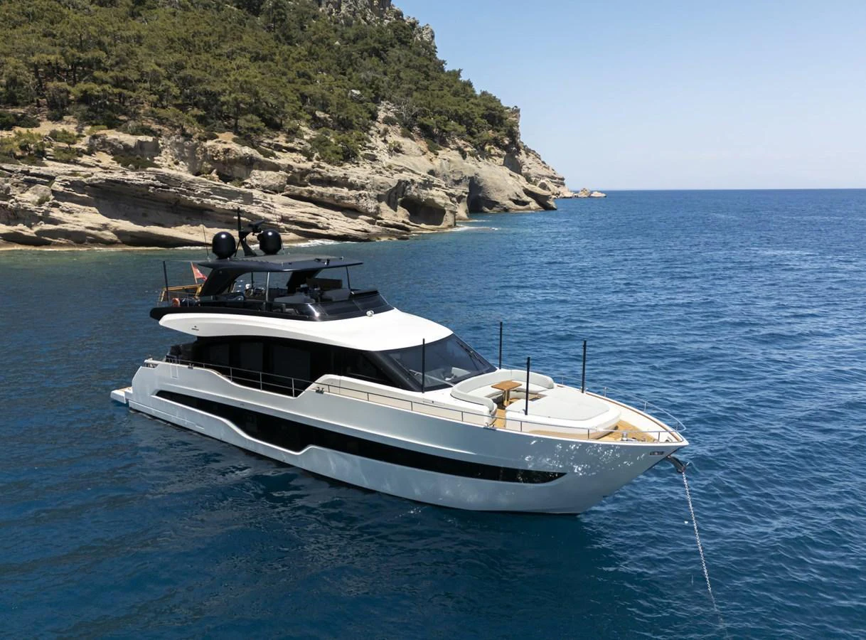 A sleek and modern yacht is anchored in a serene coastal area with clear blue waters and rocky cliffs in the background. aboard ORCA I Yacht for Sale