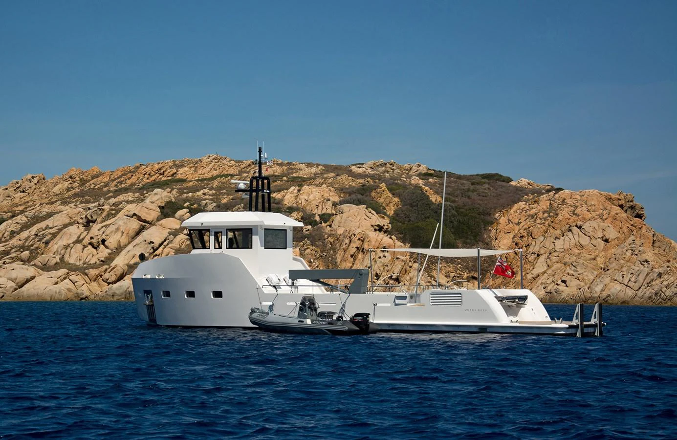 A sleek yacht is anchored near a rocky shoreline, showcasing its modern design and accompanying tender. aboard 20 YXT Yacht for Sale