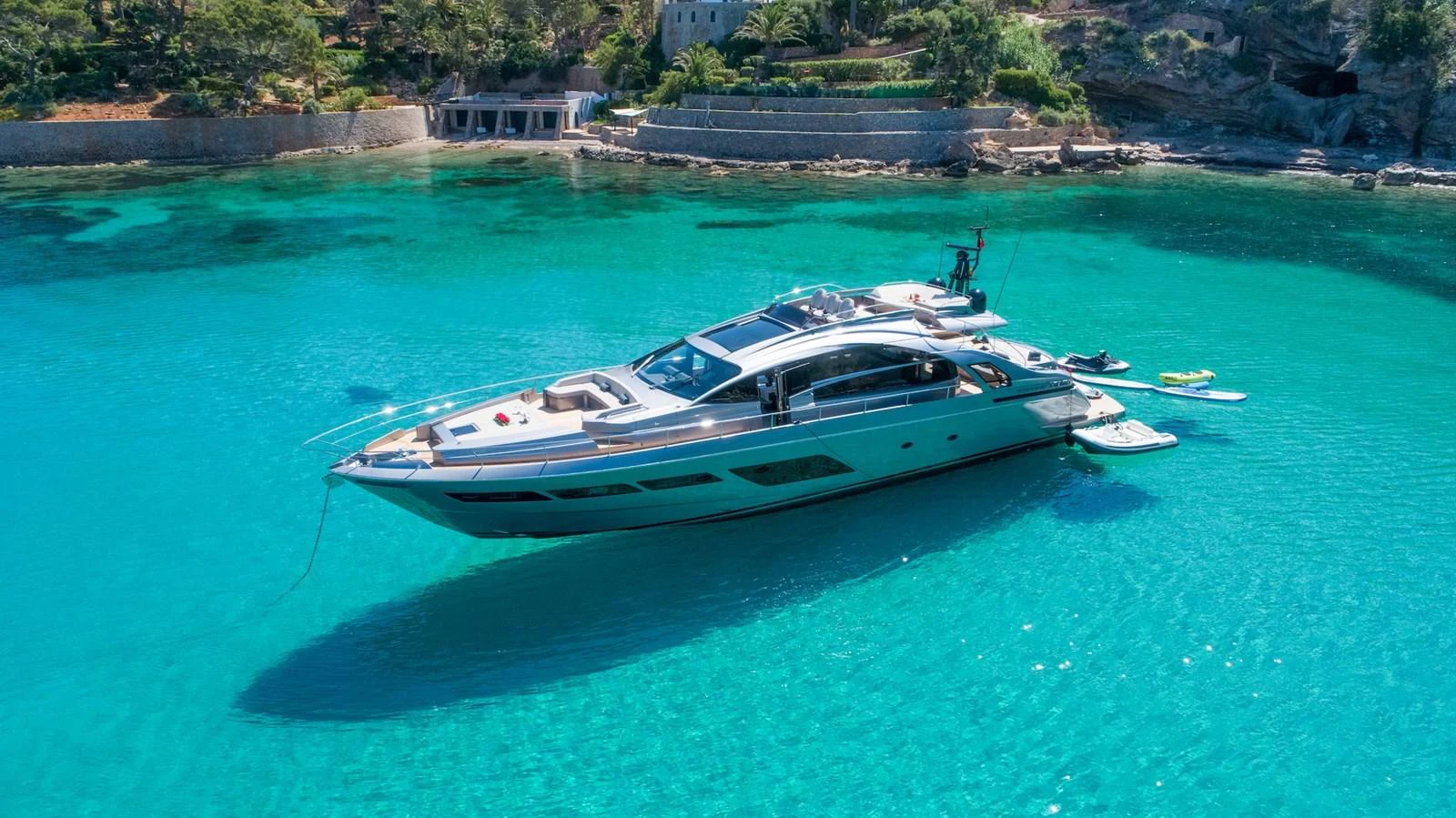 A sleek yacht is anchored in crystal-clear turquoise waters near a rocky shoreline with lush greenery. aboard DESERT STAR Yacht for Sale