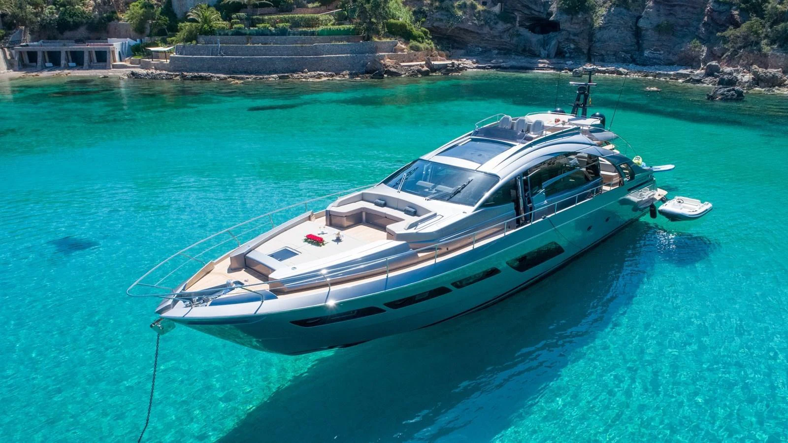 A sleek and modern yacht is anchored in crystal-clear turquoise waters near a rocky shoreline. aboard DESERT STAR Yacht for Sale