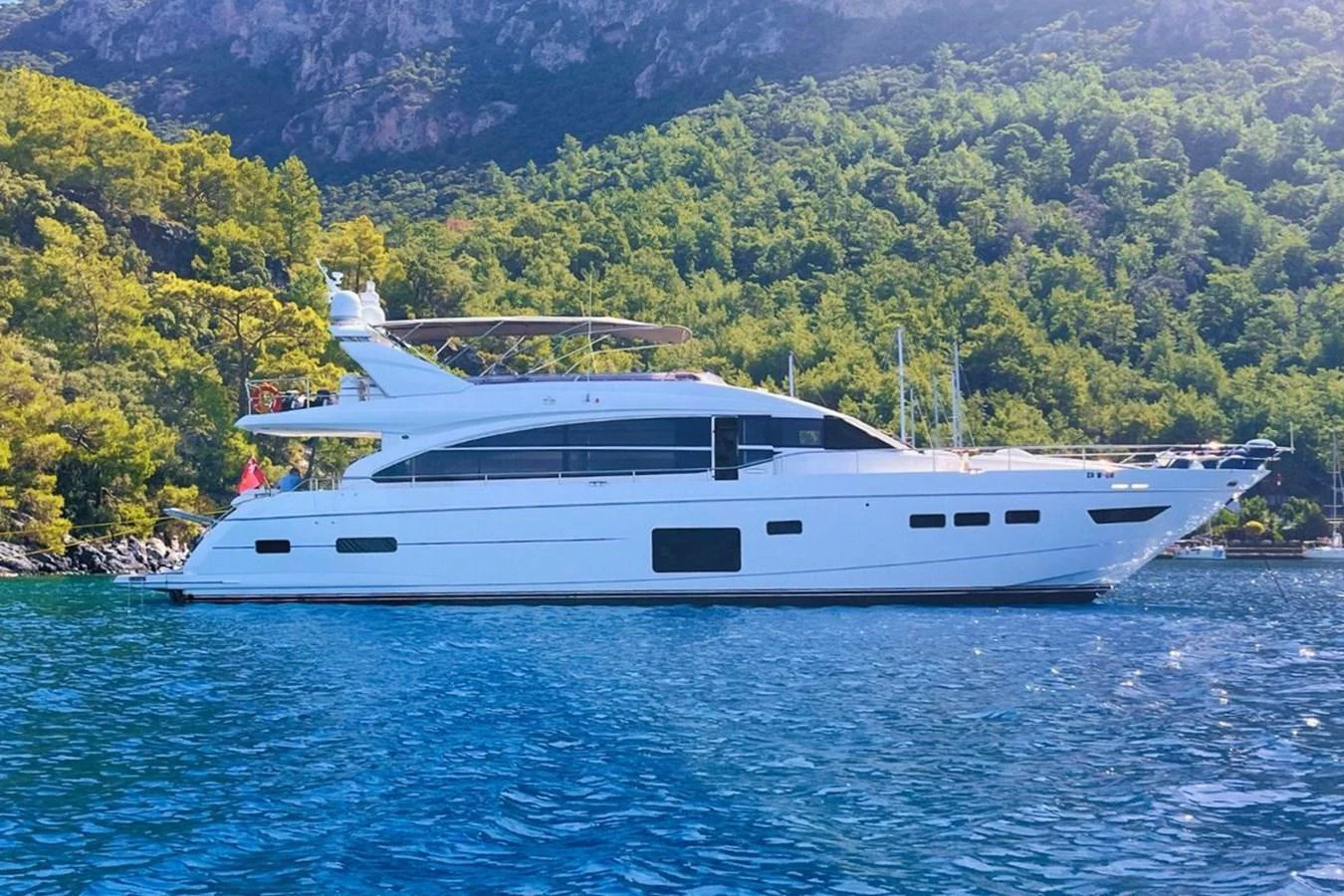 A sleek white yacht is anchored in a serene, forested bay with clear blue waters. aboard MS FELICE Yacht for Sale