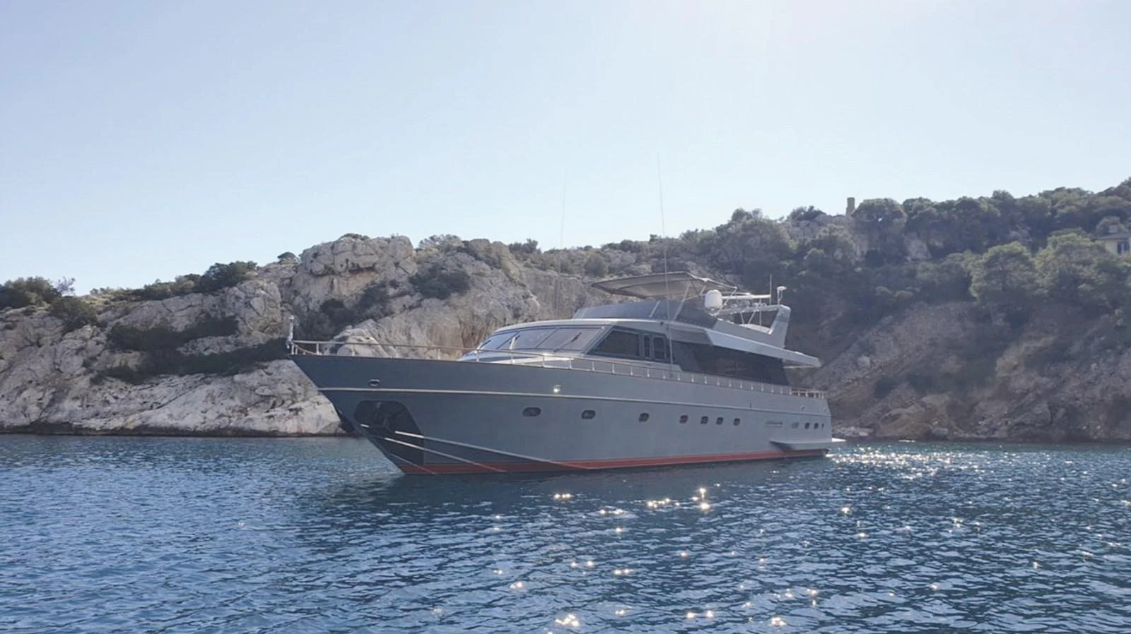 A sleek yacht is anchored in a serene bay with rocky cliffs in the background. aboard KISS Yacht for Sale