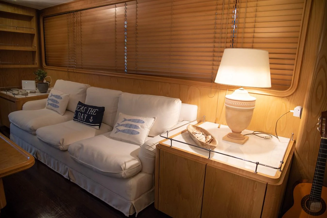 The cozy interior features a comfortable sofa with nautical-themed pillows, a warm lamp, and wooden accents. aboard KISS Yacht for Sale