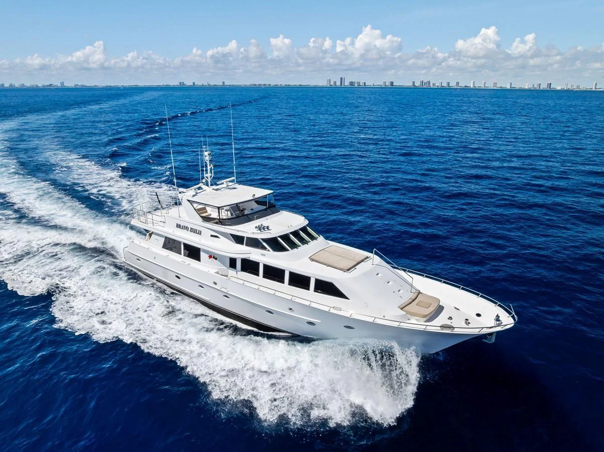 A sleek yacht cruising through the open ocean with a city skyline in the background. aboard BRAVO ZULU Yacht for Sale