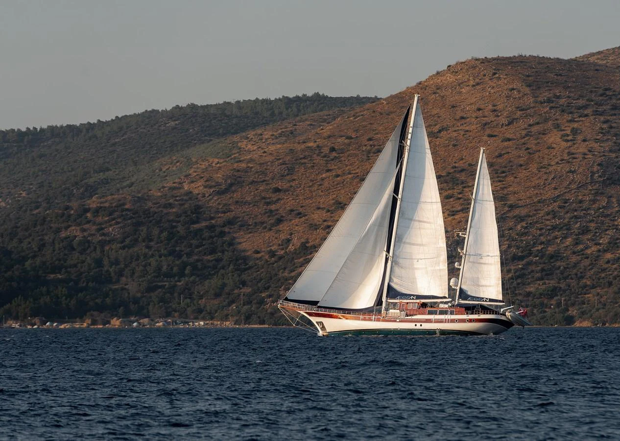 A stunning sailing yacht glides gracefully on the water against a backdrop of rugged hills. aboard AEGEAN Yacht for Sale