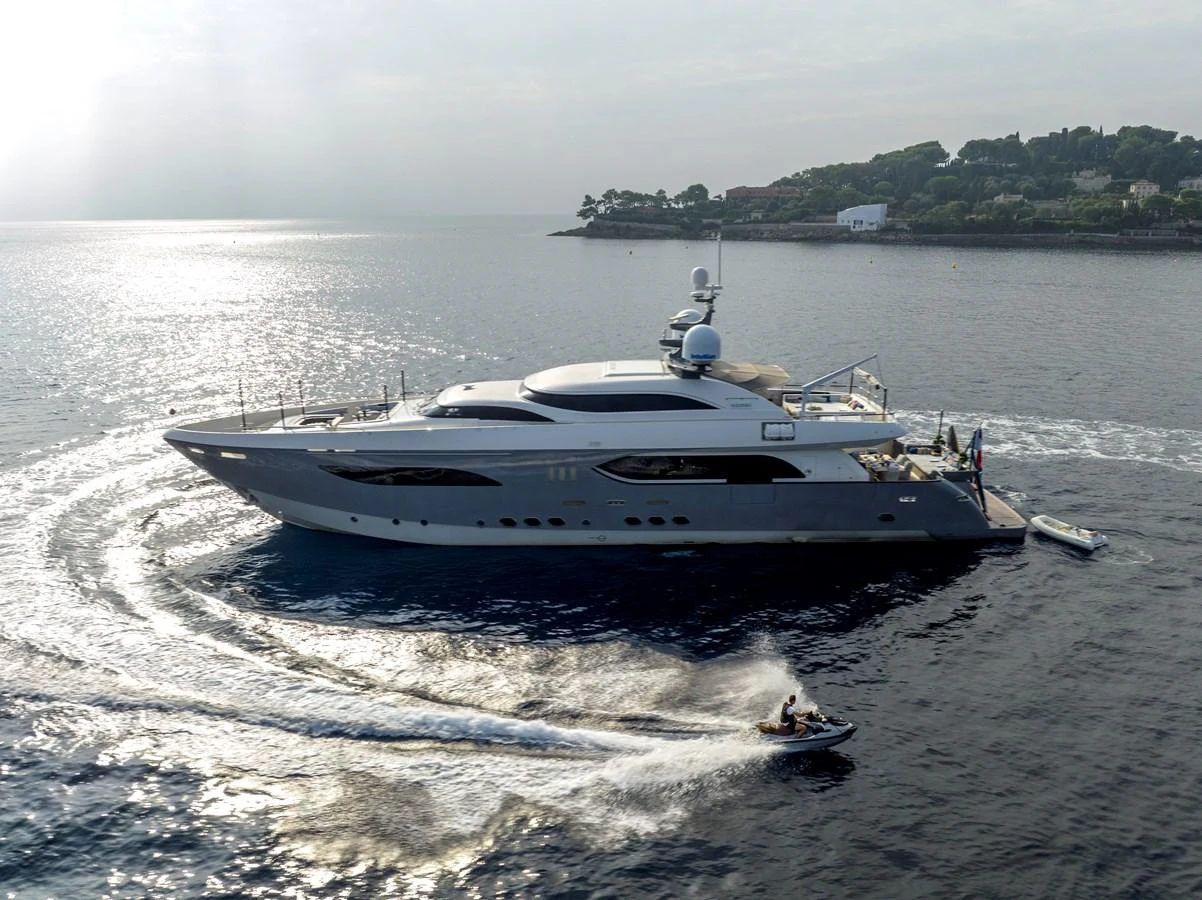 A sleek yacht cruises gracefully on calm waters, accompanied by a jet ski creating dynamic waves. aboard GEMS II Yacht for Sale