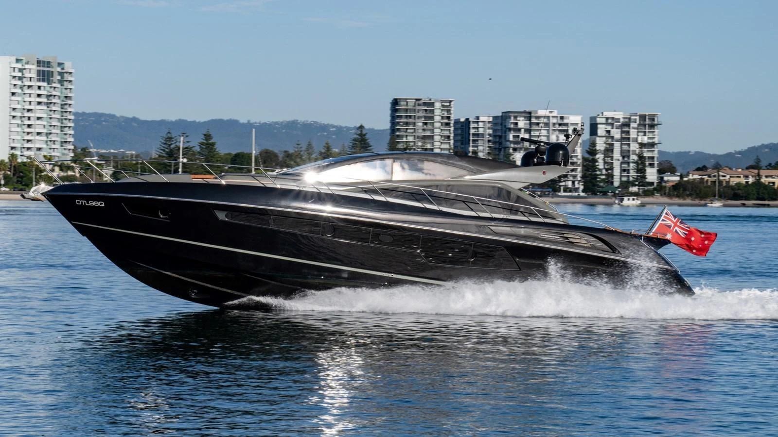 A sleek, modern yacht cruises through calm waters with a cityscape in the background. aboard ZAZA Yacht for Sale
