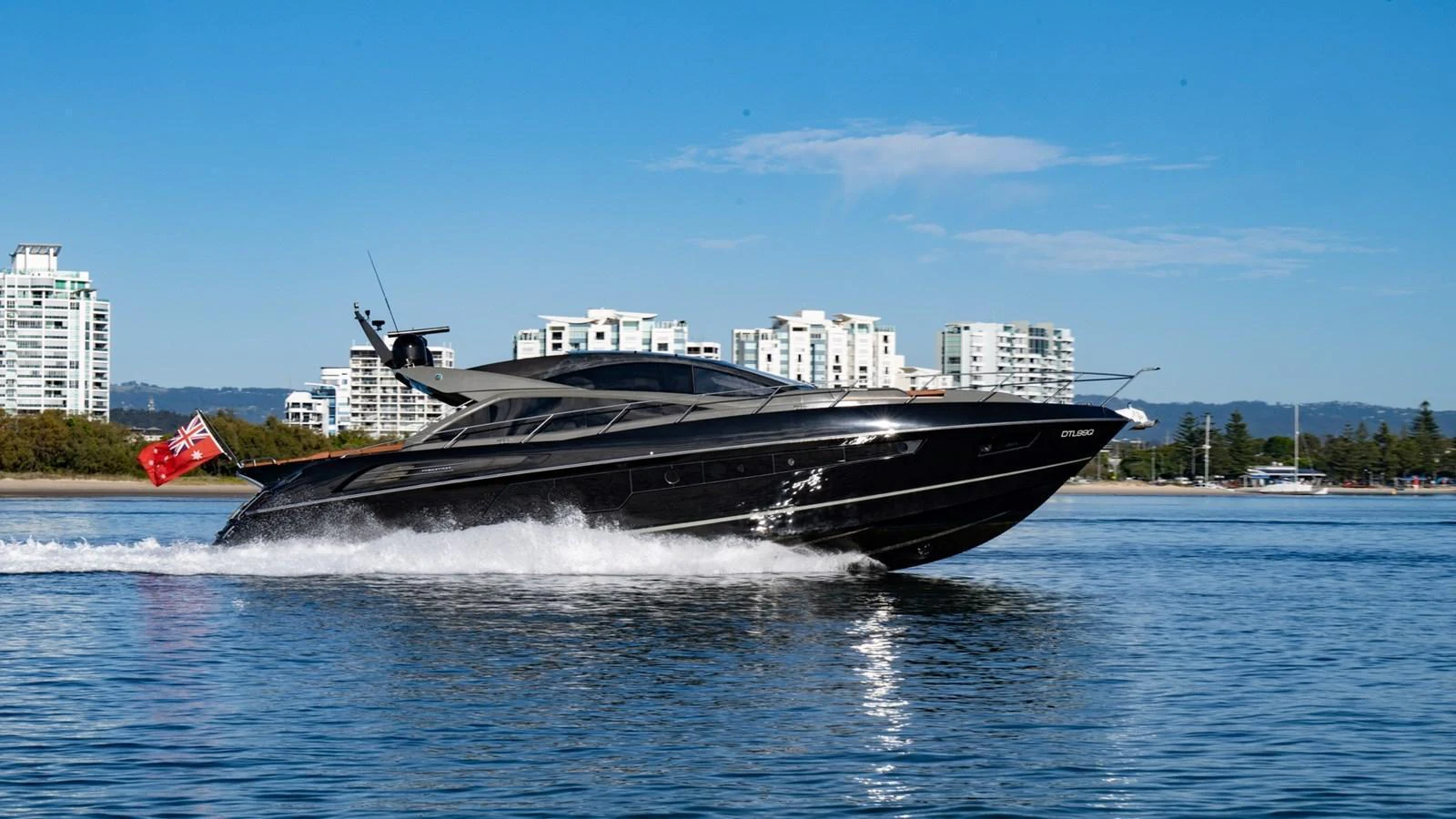 A sleek black yacht cruises swiftly on calm waters against a backdrop of modern buildings. aboard ZAZA Yacht for Sale