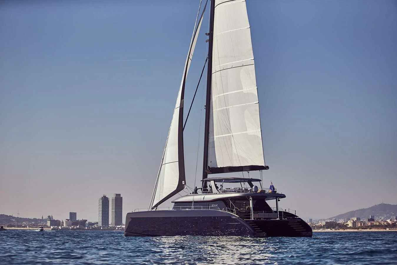 A sleek sailing yacht glides gracefully on calm waters with a cityscape in the background. aboard SUNREEF 80 NEXT Yacht for Sale