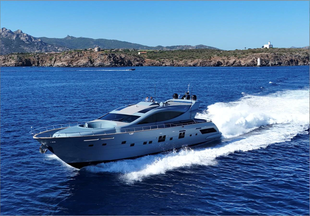 A sleek yacht cruises swiftly through clear blue waters with a scenic coastline in the background. aboard DEEA II Yacht for Sale