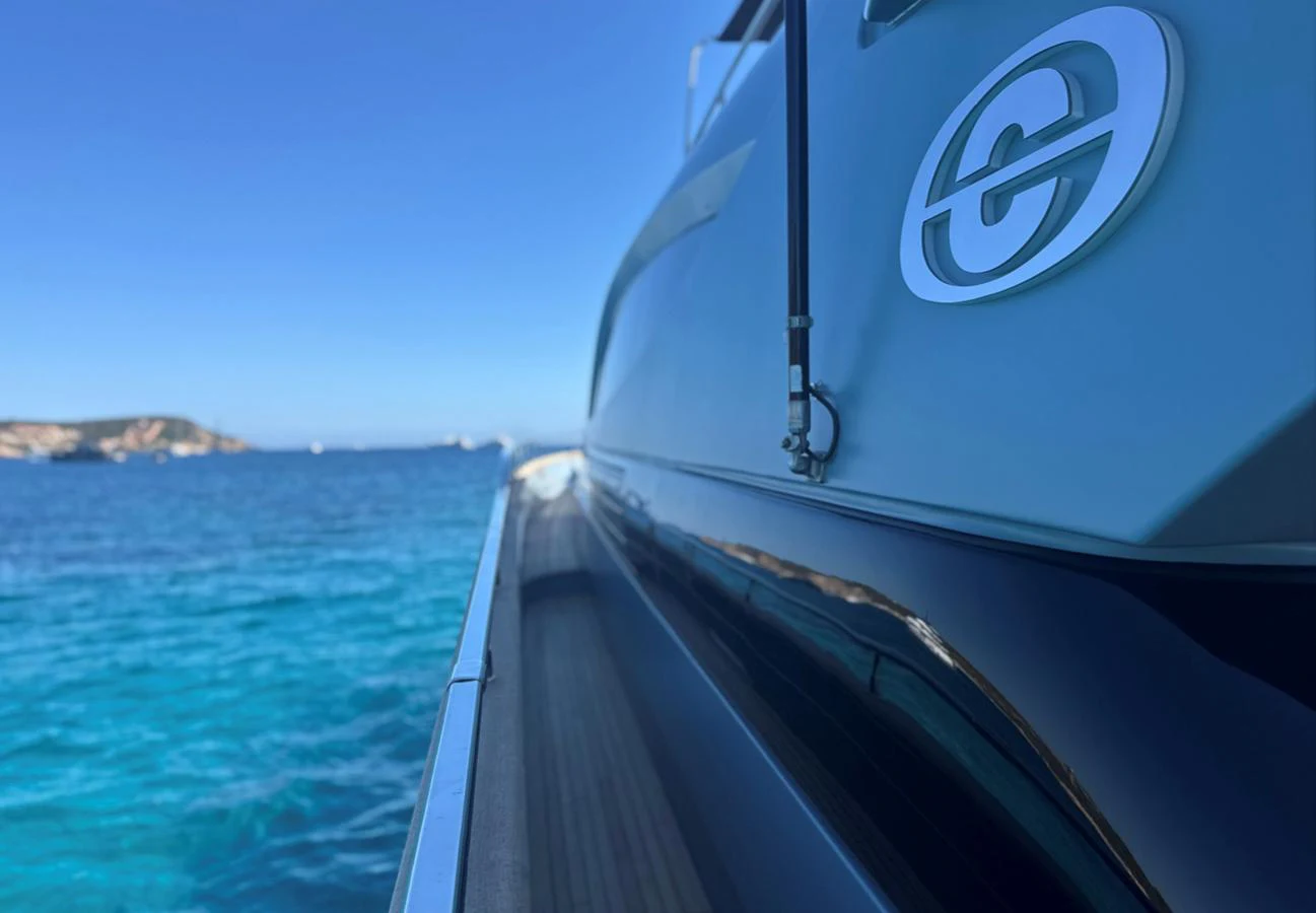 A close-up view of the yacht's sleek exterior with a logo, set against a backdrop of clear blue waters. aboard DEEA II Yacht for Sale