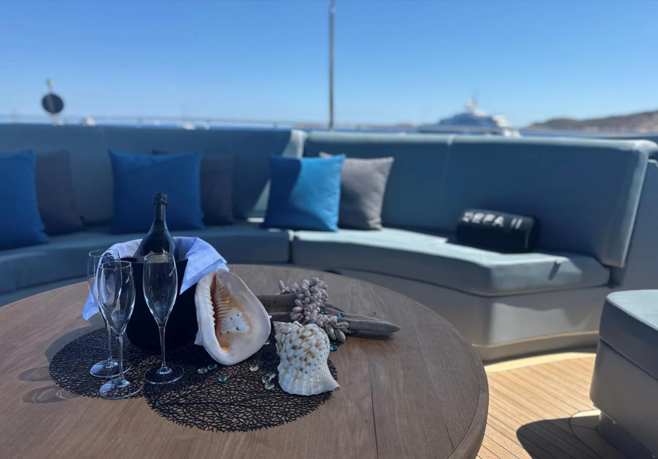 An elegant outdoor lounge area with a wooden table adorned with seashells, champagne, and glasses, set against a backdrop of the sea. aboard DEEA II Yacht for Sale