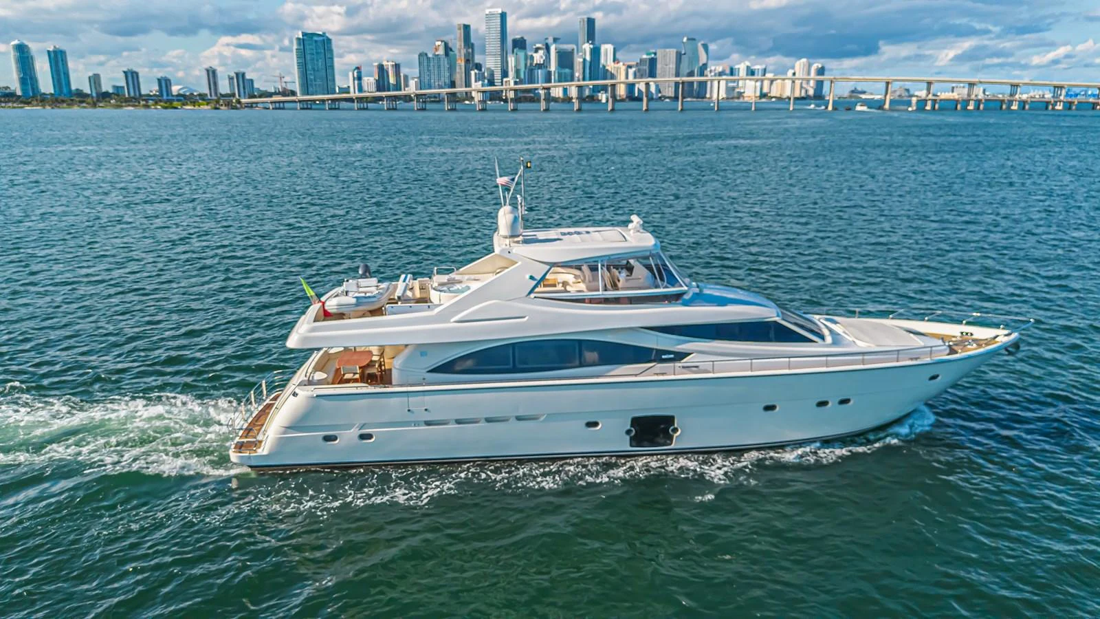 A sleek yacht cruises through calm waters with a cityscape in the background. aboard NORAL Yacht for Sale