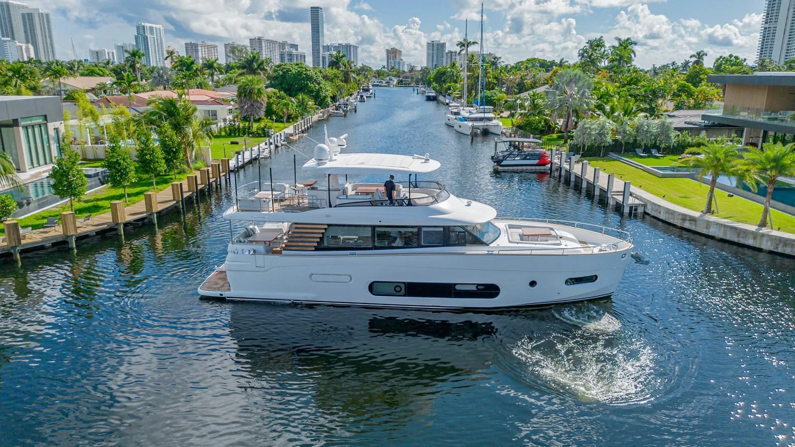 A sleek yacht cruises through a serene canal lined with lush greenery and modern buildings. aboard XAX66M61L122 Yacht for Sale