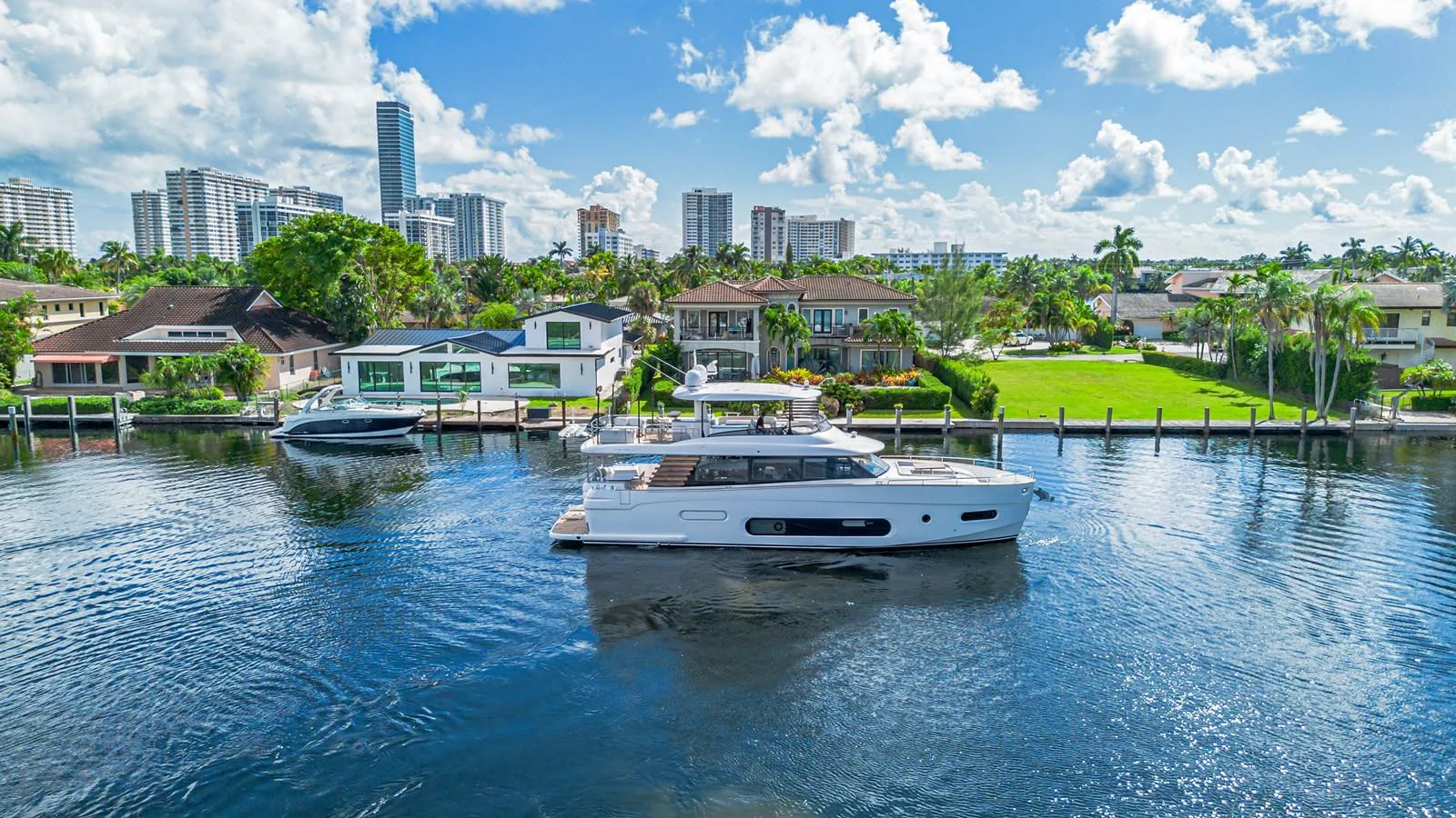 A sleek yacht cruises along a serene waterfront with a backdrop of luxurious homes and city skyline. aboard XAX66M61L122 Yacht for Sale