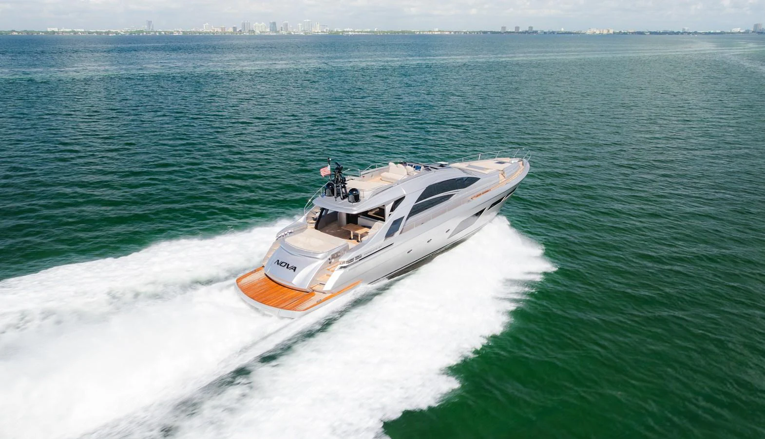 A sleek yacht cruises swiftly across calm waters with a cityscape in the background. aboard NOVA Yacht for Sale