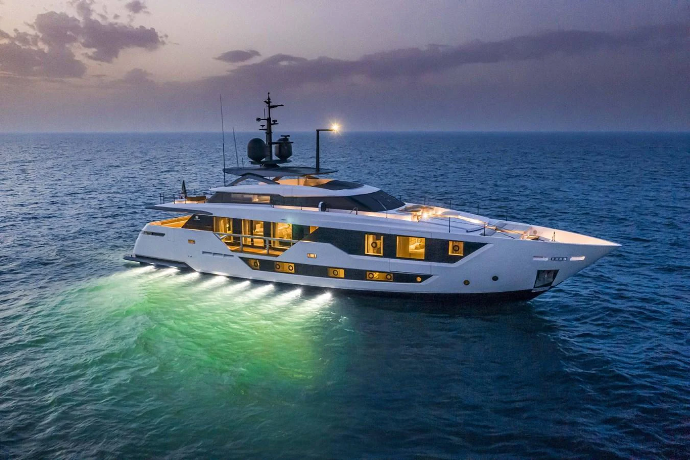 A sleek and modern yacht cruises through the open sea at dusk, illuminated by underwater lights. aboard CUSTOM LINE SAETTA 128/02 Yacht for Sale