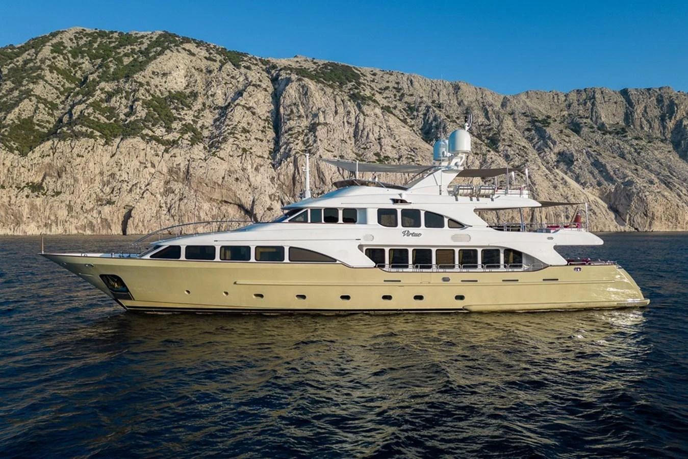 A luxurious yacht cruises along a rocky coastline under a clear blue sky. aboard VIRTUE Yacht for Sale