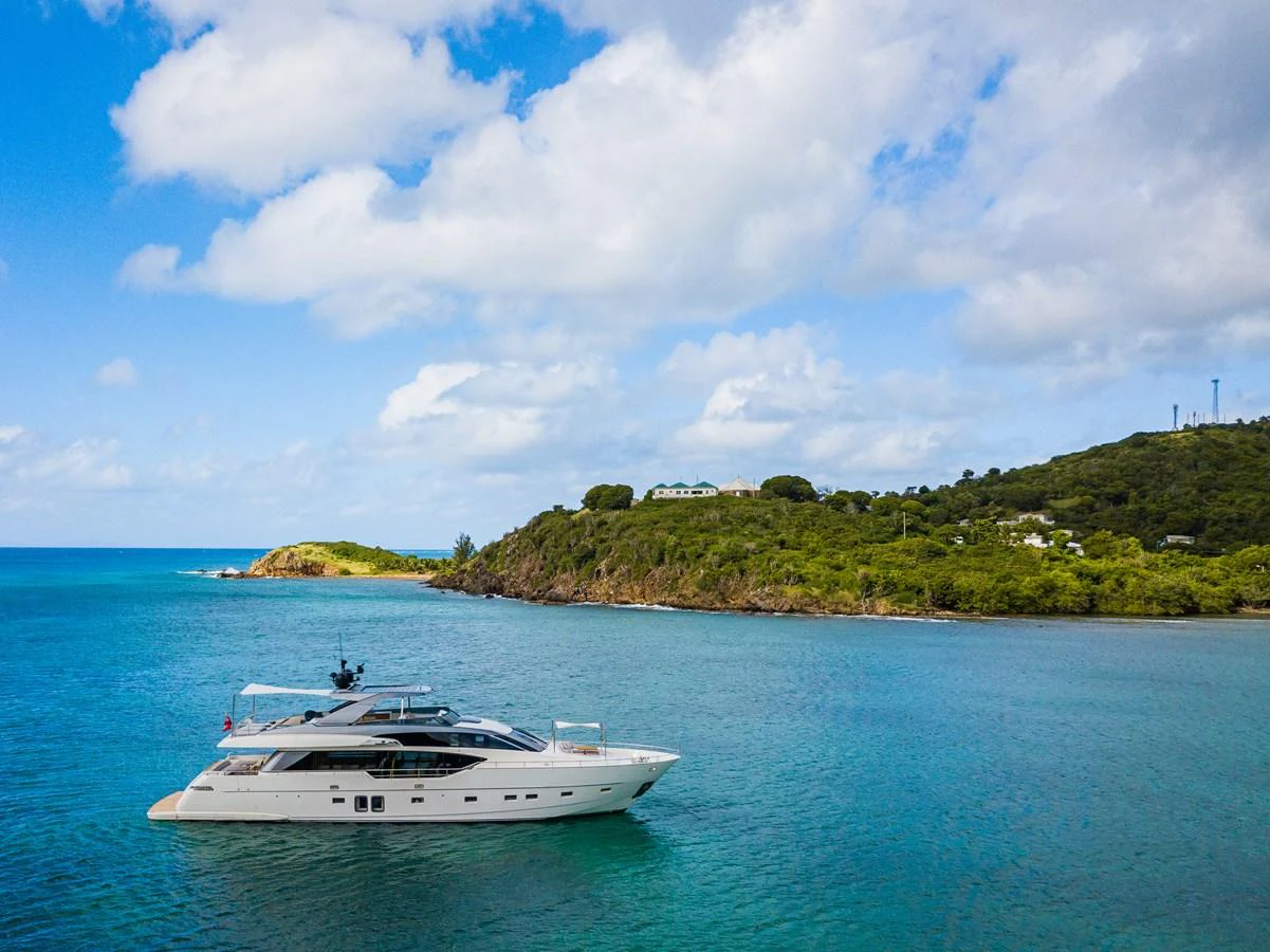 A sleek yacht cruises through tranquil blue waters against a backdrop of lush green hills and a partly cloudy sky. aboard AKULA Yacht for Sale