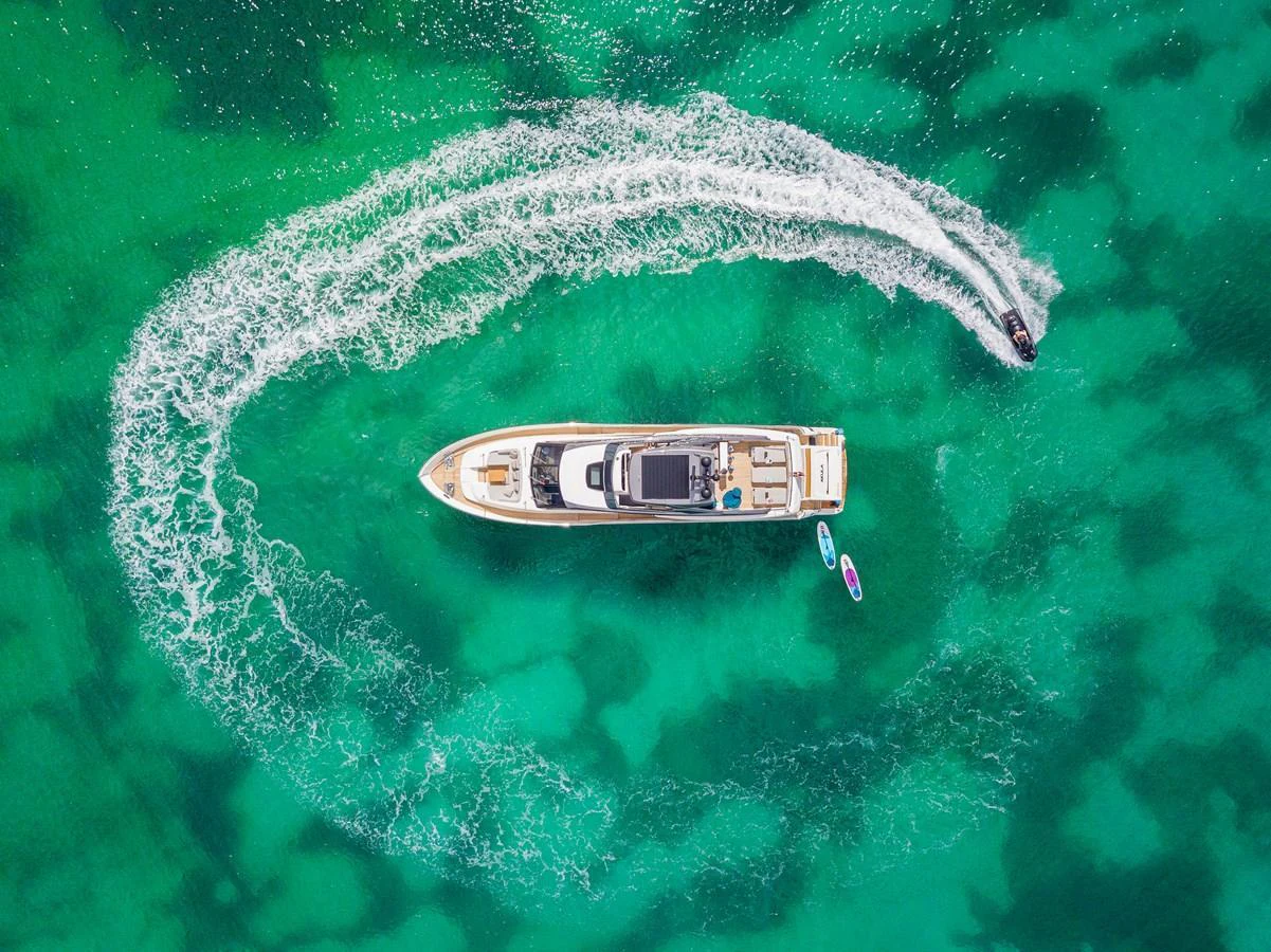 A stunning aerial view of a yacht cruising in clear turquoise waters, with a jet ski creating a circular wake around it. aboard AKULA Yacht for Sale